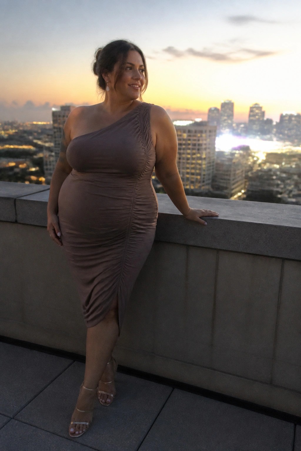 Woman in taupe one-shoulder ruched dress leans on balcony overlooking city at dusk