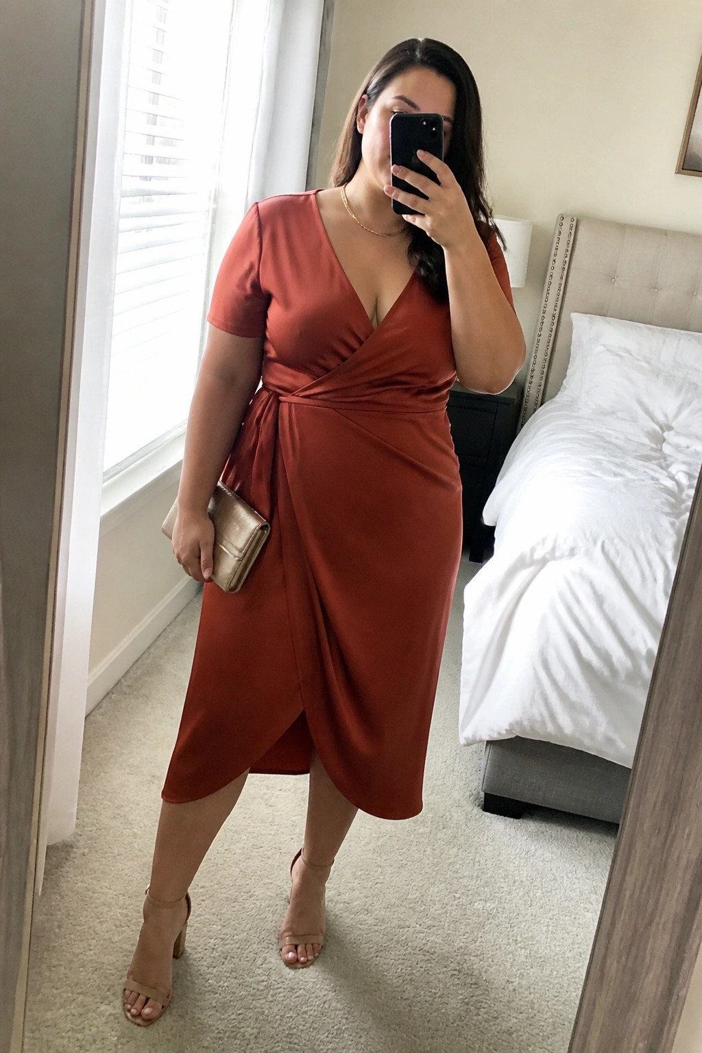 Plus size woman wearing a rust orange satin wrap midi dress with short sleeves, paired with nude block heel sandals and a gold clutch, taking a mirror selfie in a bedroom setting.