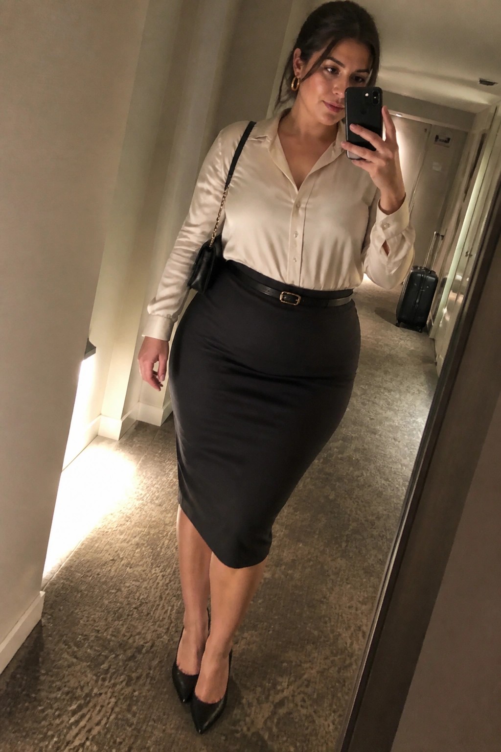 Woman in cream satin blouse and black pencil skirt taking mirror selfie in hallway
