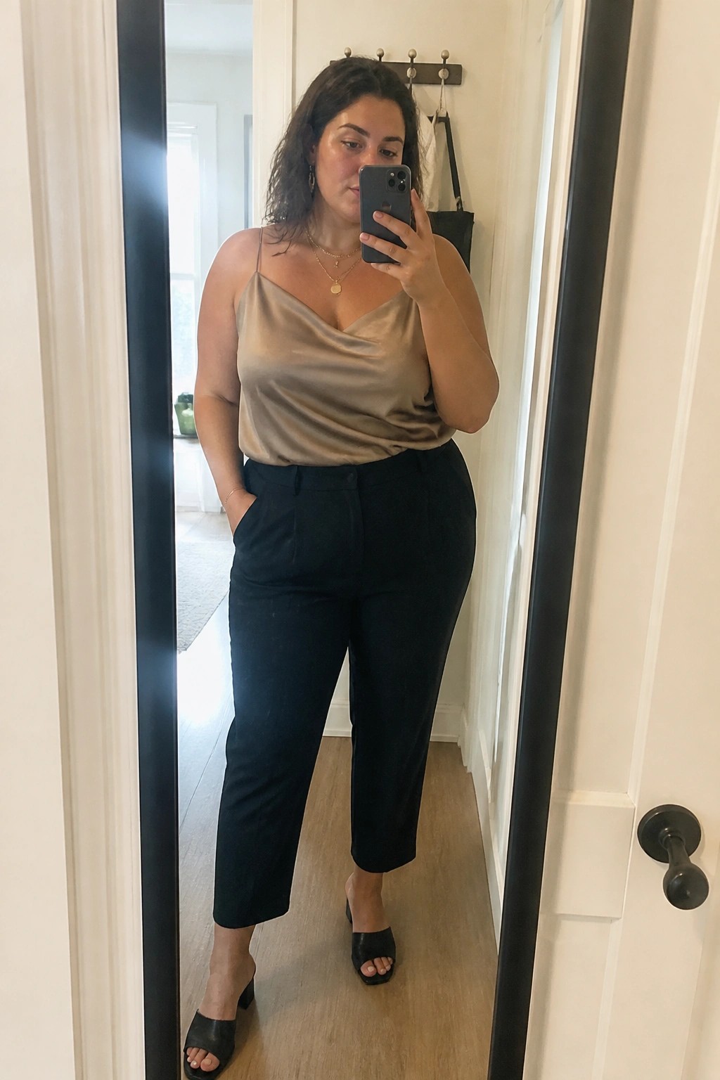 Plus size woman in a beige satin cowl neck camisole and black cropped trousers with black heeled mules taking a mirror selfie in a hallway