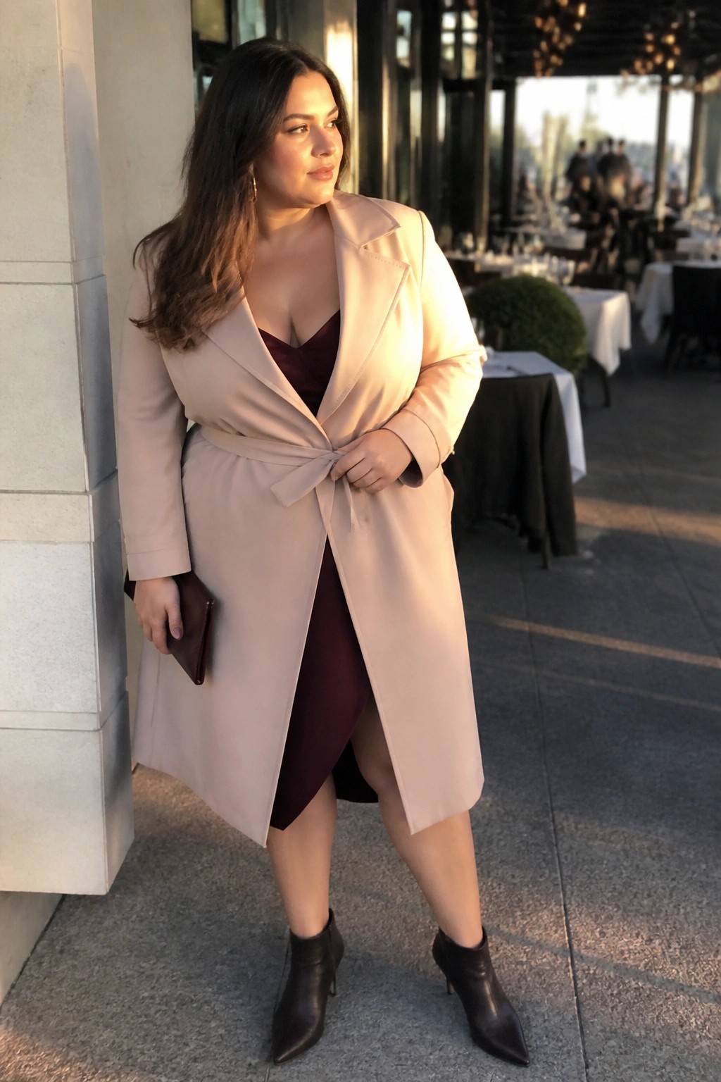 Woman in beige belted coat over burgundy dress holding clutch at outdoor restaurant.