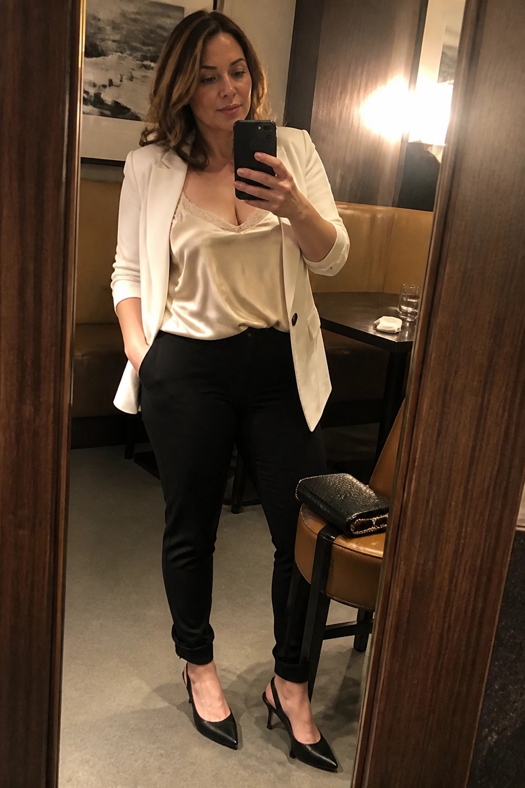 Woman in white blazer and black pants taking mirror selfie in restaurant.