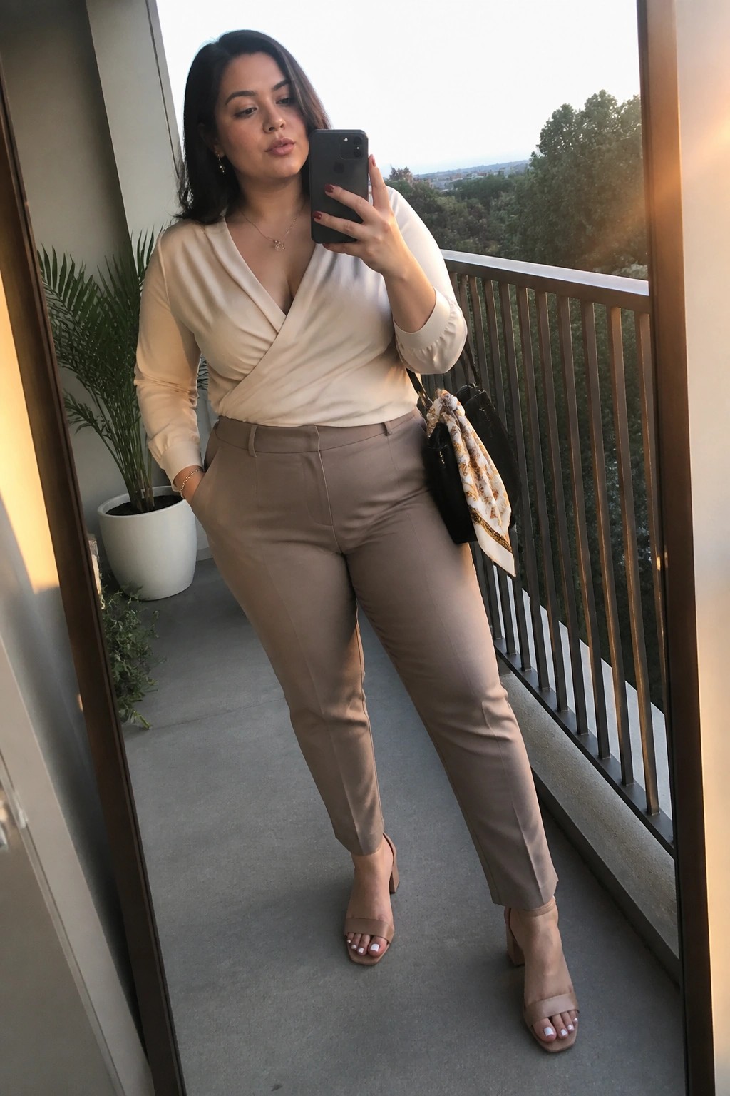 Plus size woman wearing a cream wrap blouse tucked into taupe tailored pants with nude block heel sandals and a black crossbody bag.