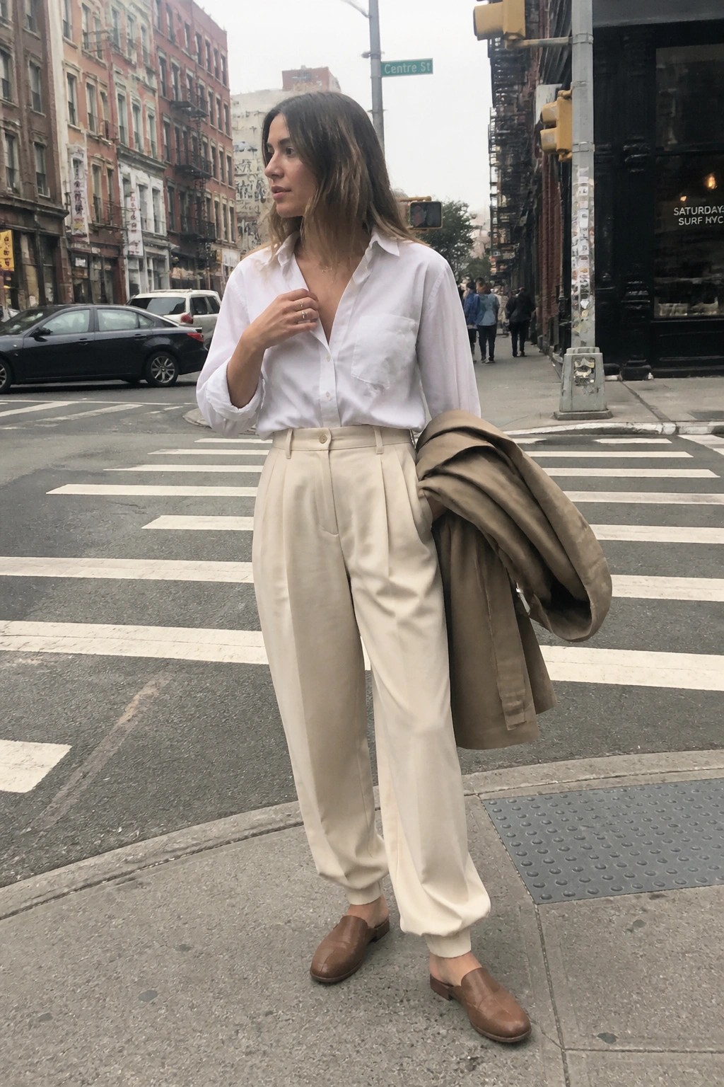 Woman in white shirt and beige trousers holding coat on city crosswalk.