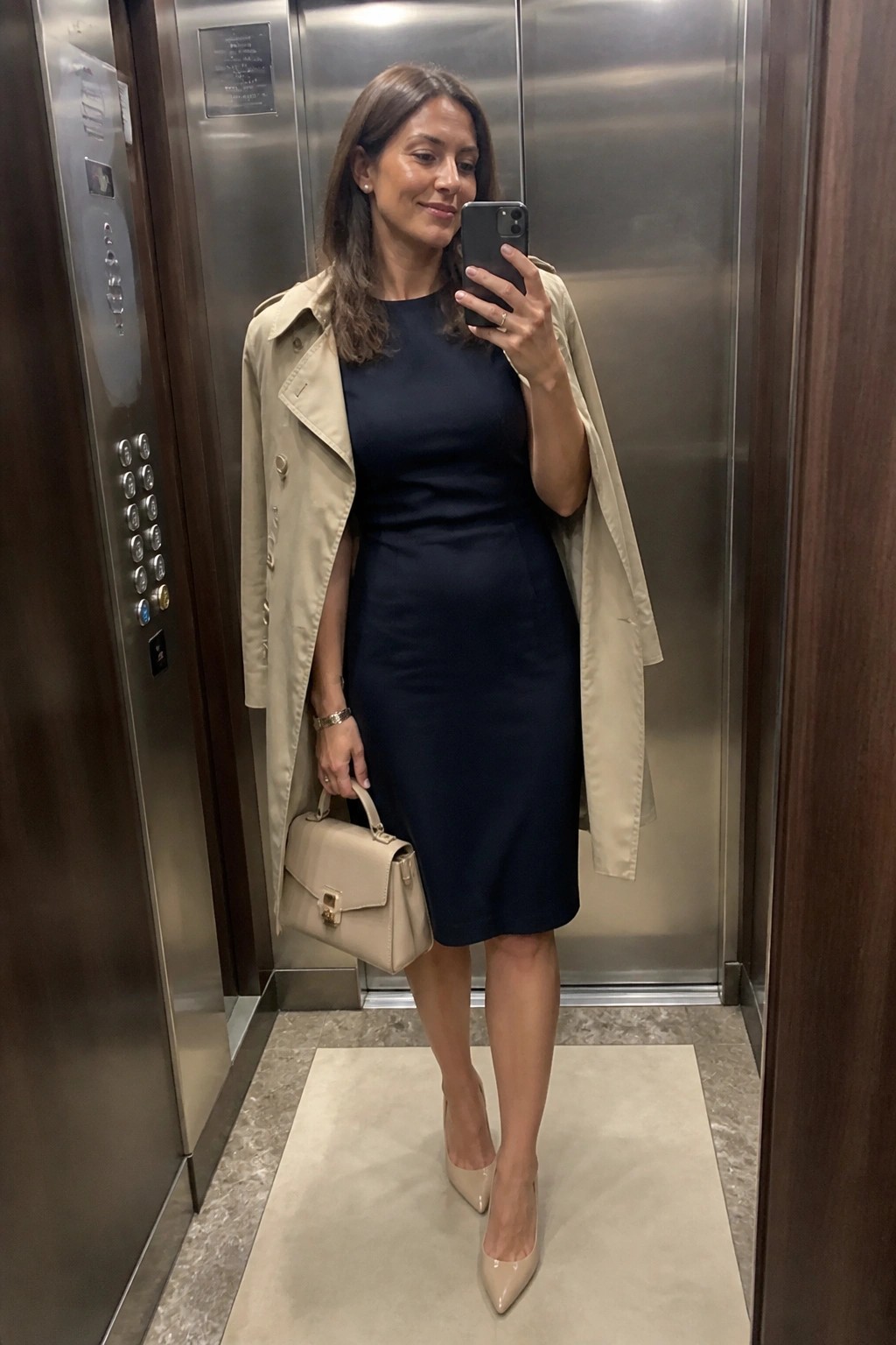 Woman wearing a navy knee-length sheath dress with a beige trench coat draped over her shoulders, holding a matching beige handbag and phone, paired with beige pointed-toe heels in an elevator setting.