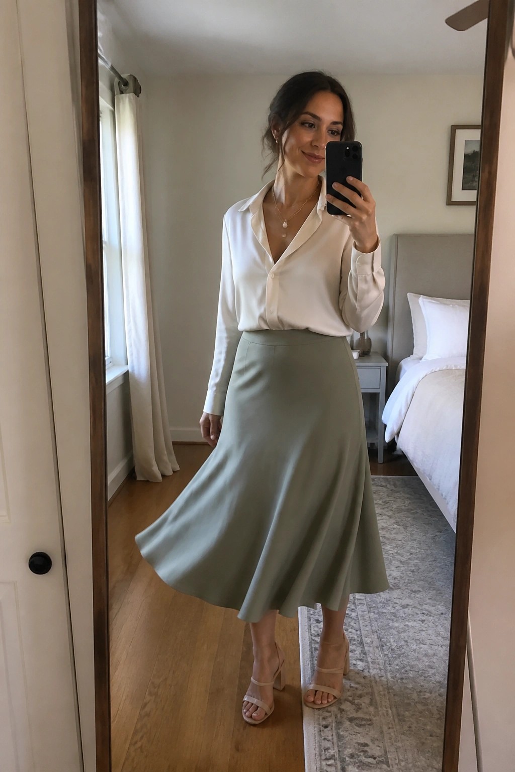 Woman taking a mirror selfie in a white satin button-down blouse tucked into a sage green midi skirt, paired with beige strappy block heels and a delicate gold necklace with a pearl pendant.