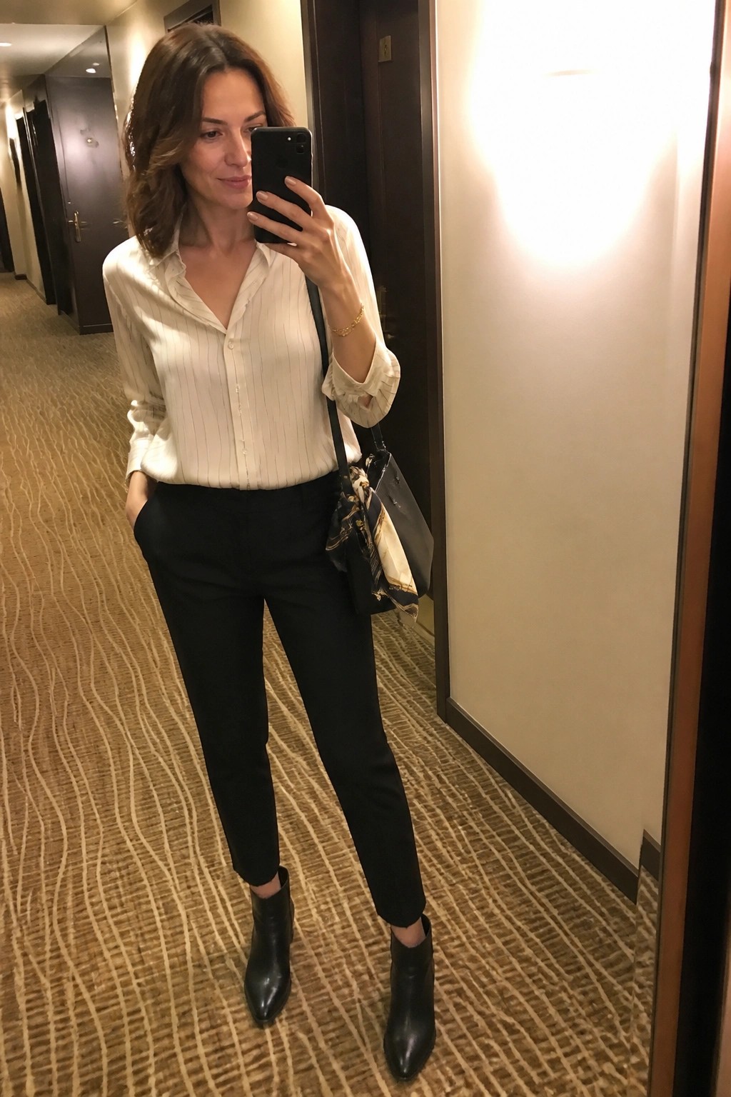 Woman in white striped shirt and black pants taking mirror selfie in hallway