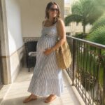 Pregnant woman in blue striped dress holding straw bag on sunny ocean balcony