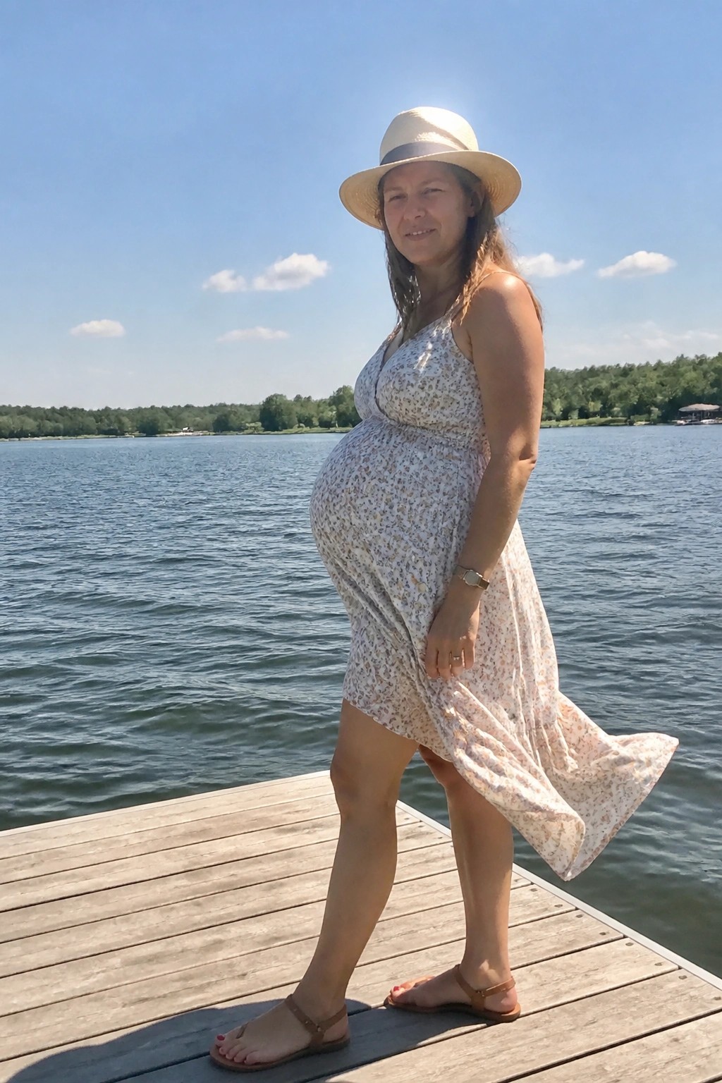 Pregnant woman wearing a light floral print maternity sundress with spaghetti straps, paired with brown sandals and a straw hat, standing on a wooden dock beside water.