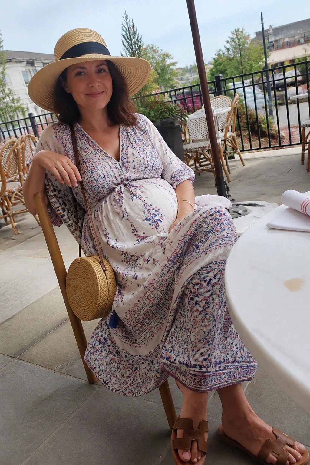 Pregnant woman in floral dress and straw hat seated at outdoor café patio