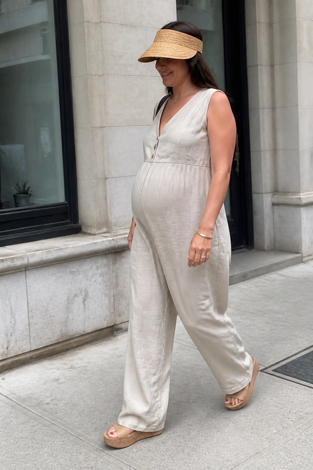 Pregnant woman in beige linen jumpsuit and straw visor walking on sidewalk