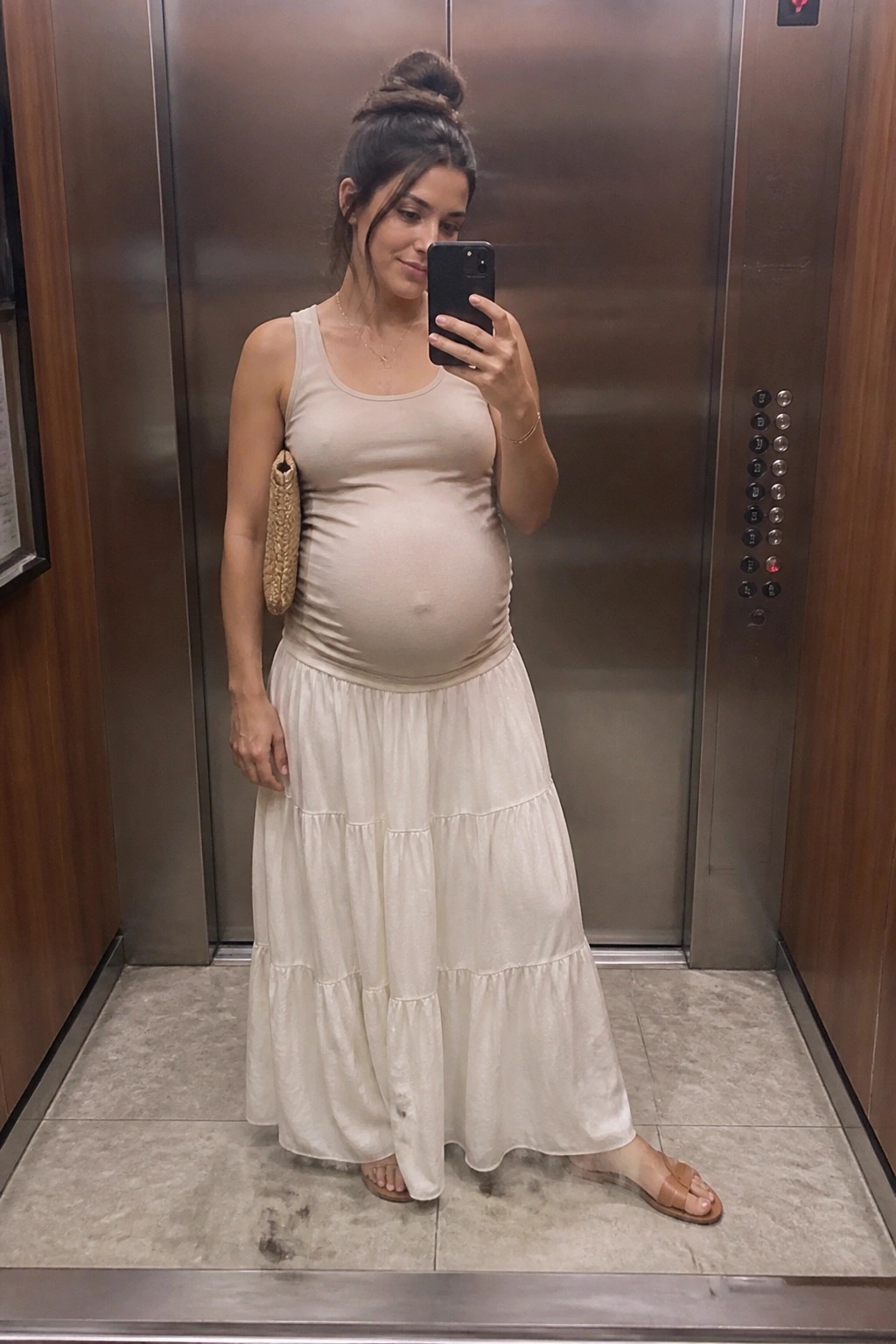 Pregnant woman in beige tank top and tiered white skirt taking elevator selfie