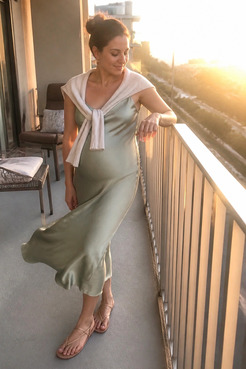 Woman in sage green satin dress with white scarf on sunset balcony