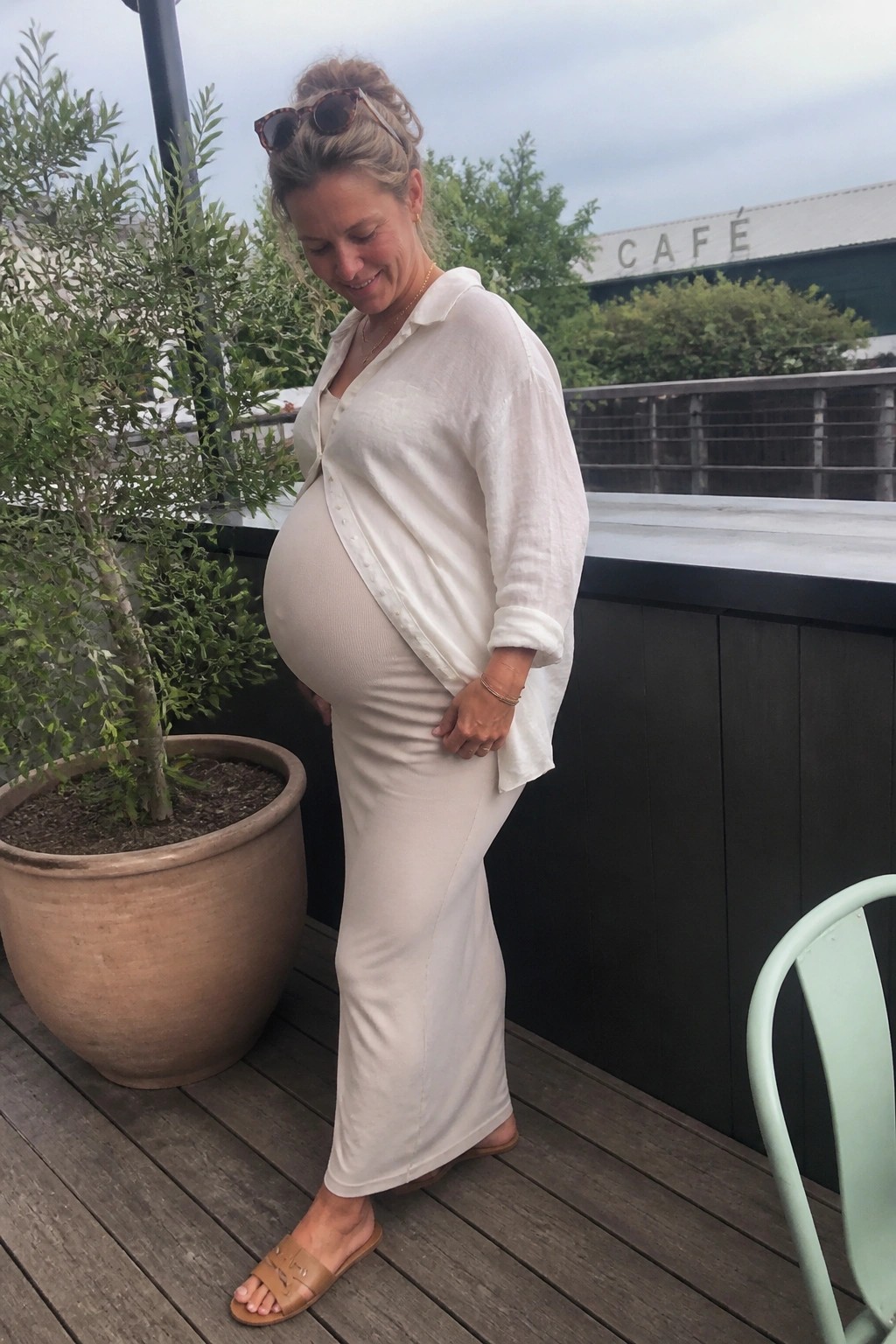 Pregnant woman in white outfit smiles while touching belly on wooden deck