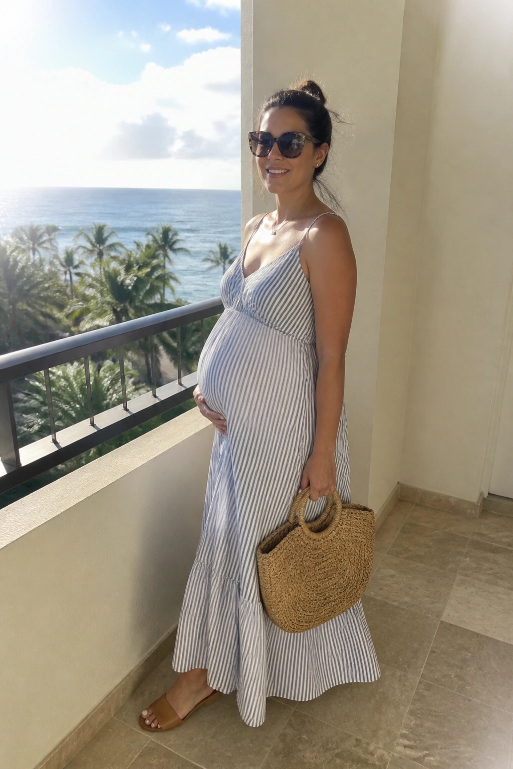 Pregnant woman in blue striped dress holding straw bag on sunny ocean balcony