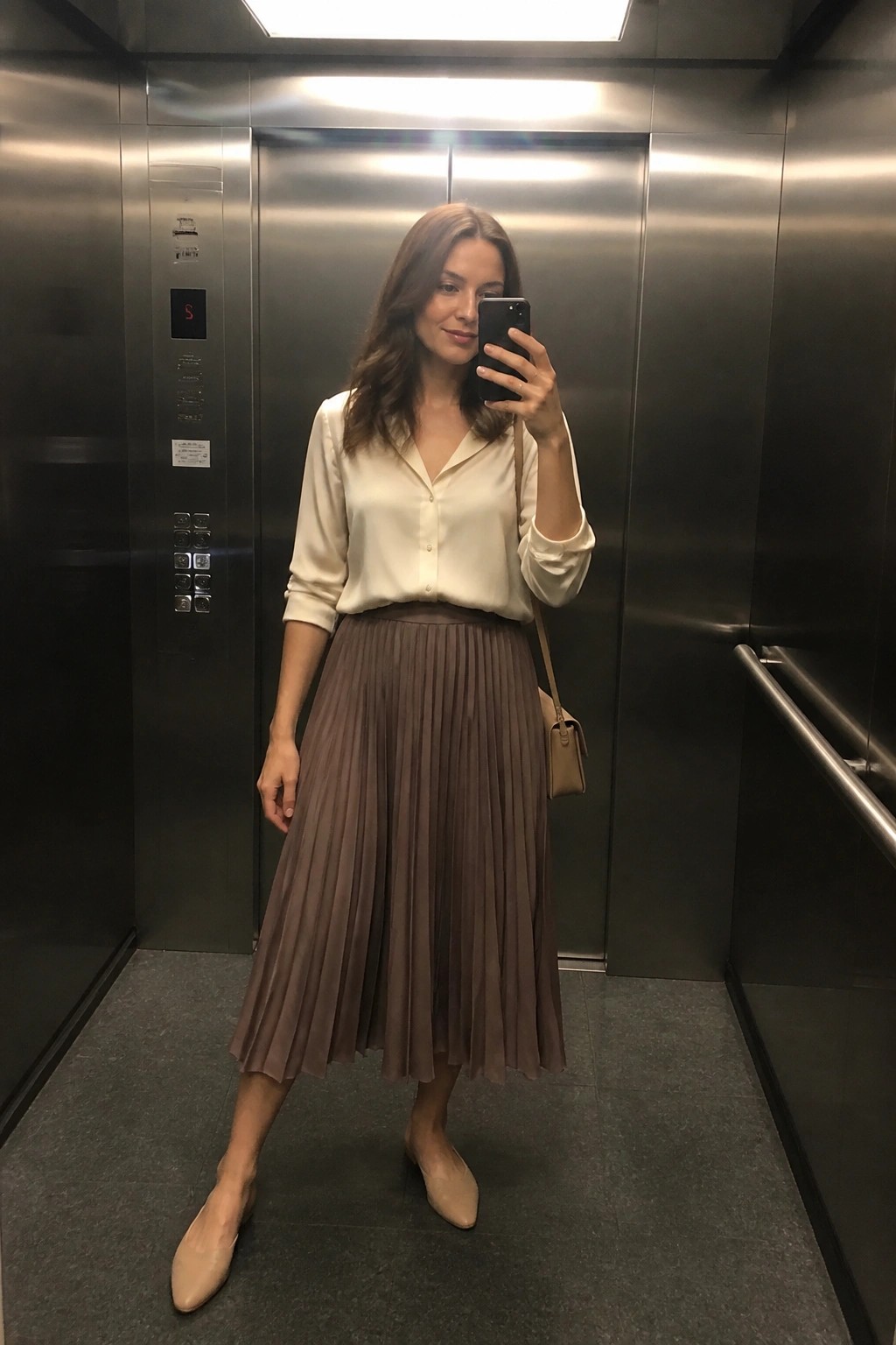 Woman standing in an elevator wearing a cream satin blouse tucked into a taupe pleated midi skirt with beige pointed flats and a small crossbody bag, holding a phone for a mirror selfie.