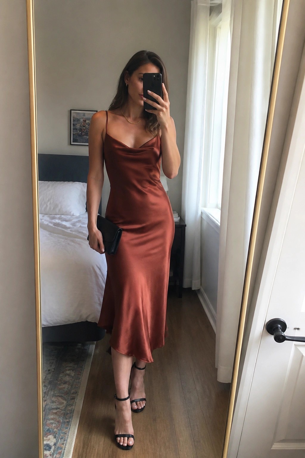 Woman in rust satin slip dress taking mirror selfie holding phone and clutch