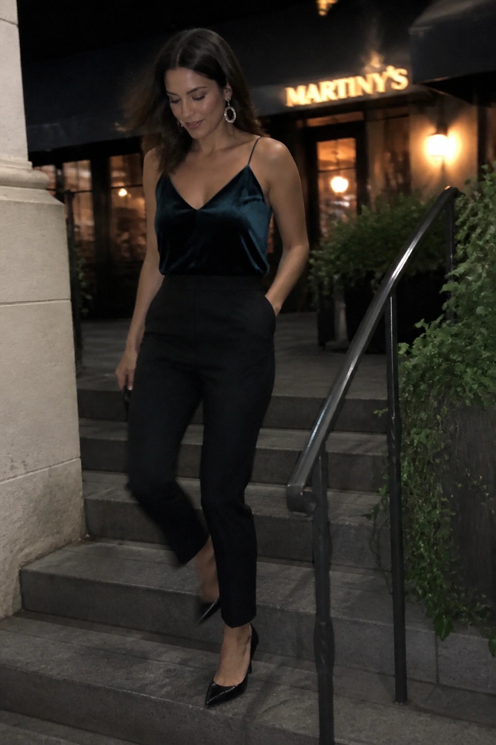 Woman in teal velvet camisole and black pants descending stone stairs at night