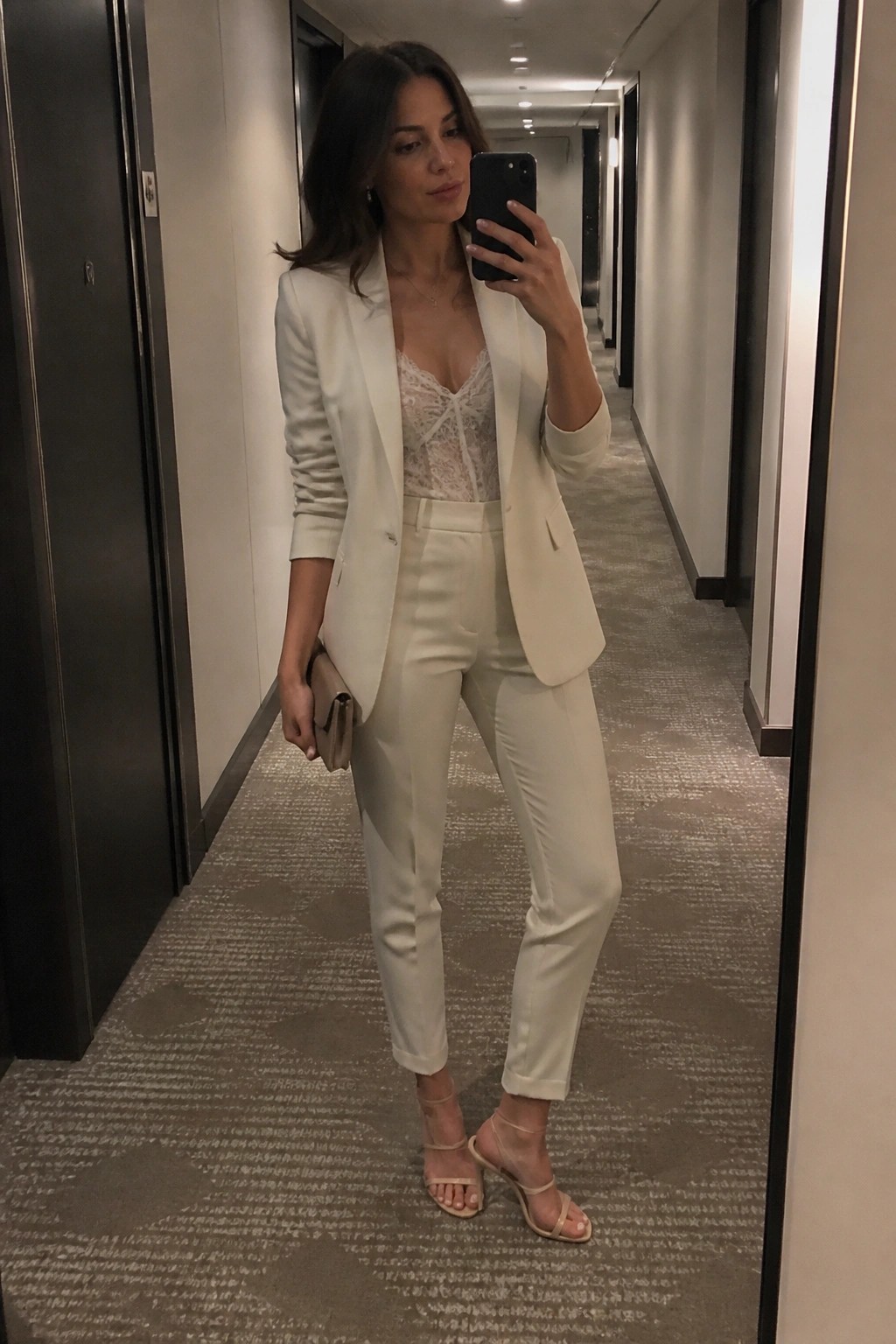 Woman in white pantsuit taking mirror selfie in hallway with phone and clutch