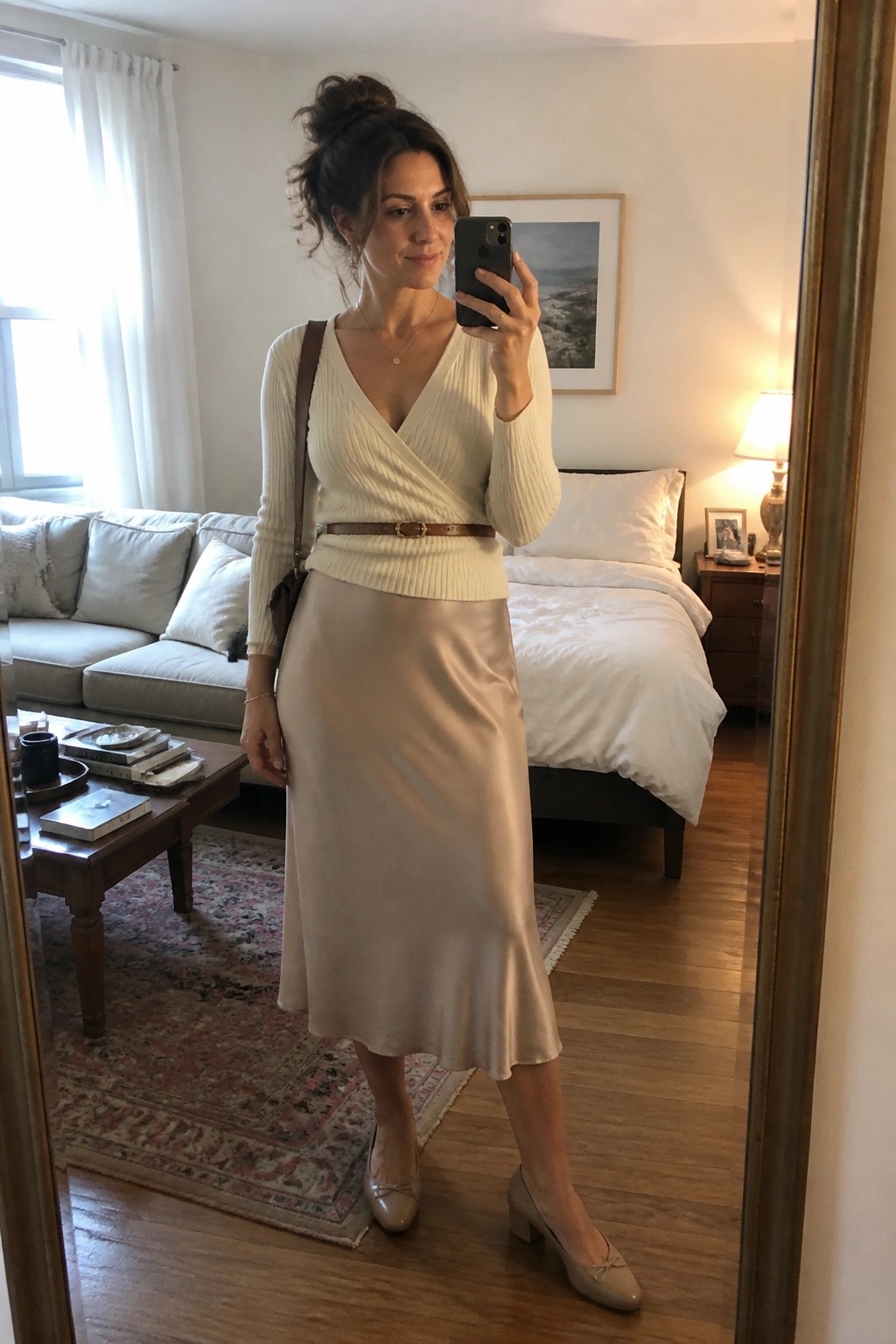 Woman taking a mirror selfie in a cream wrap sweater tucked into a beige satin midi skirt with brown belt, neutral heeled shoes, and crossbody bag in a living room setting.