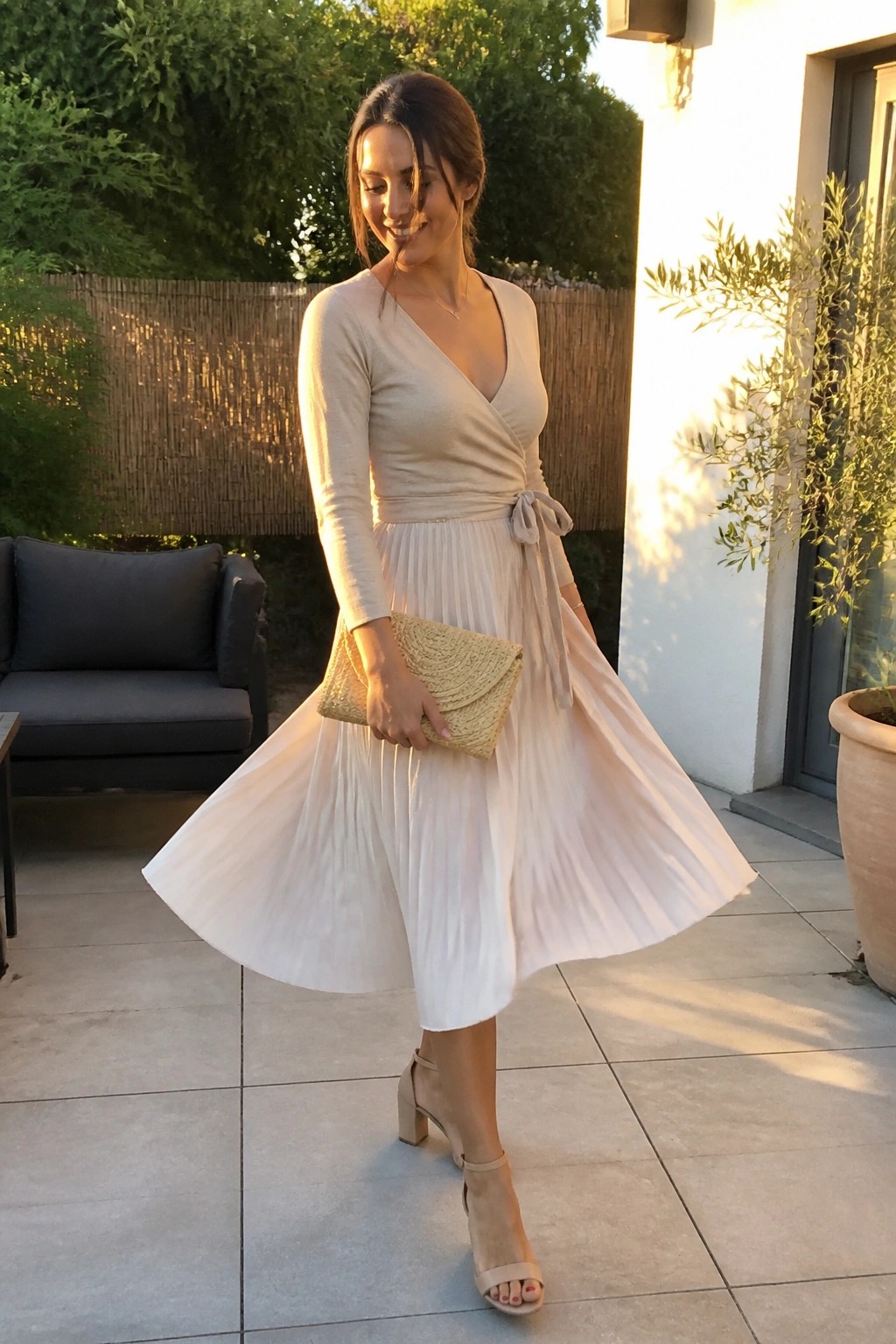 Woman in cream wrap top with side tie and long sleeves paired with white pleated midi skirt, beige block heels, and woven clutch.