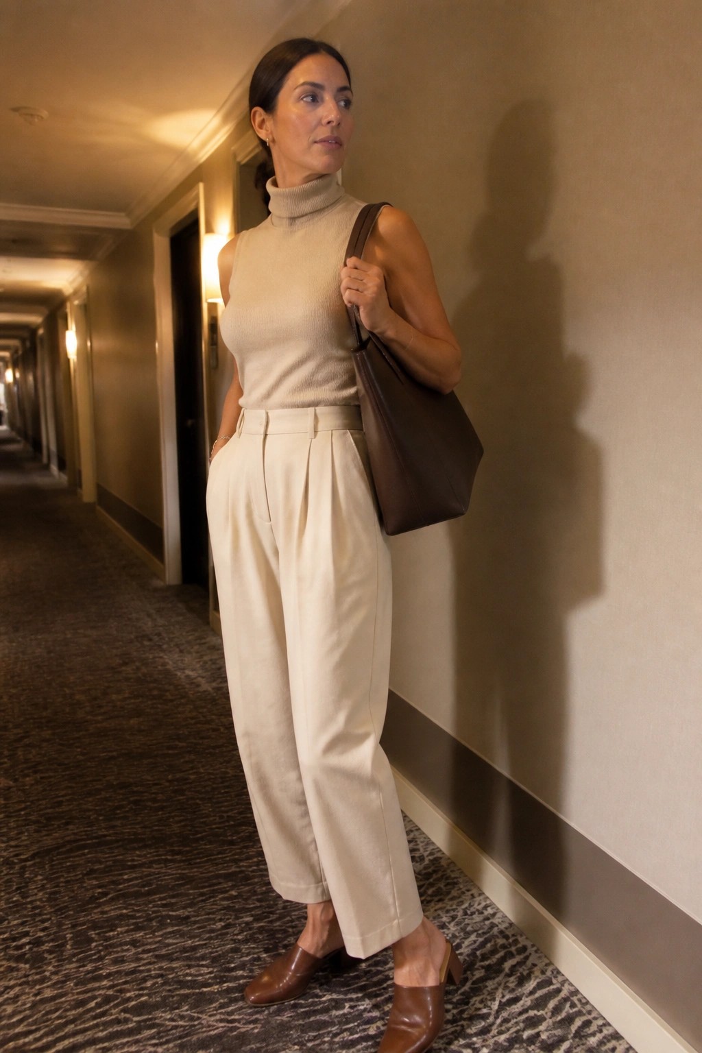 Woman in a beige sleeveless turtleneck sweater tucked into cream wide-leg pleated trousers, paired with brown leather mules and a brown leather tote bag.