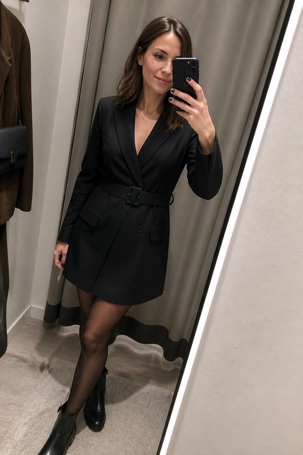 Woman wearing a black wrap blazer dress with a wide belt, sheer black tights, and black ankle boots while taking a mirror selfie in a fitting room.