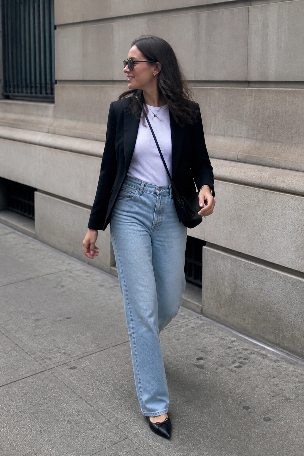Woman wearing black blazer over white top with light blue jeans, black pointed flats and crossbody bag.