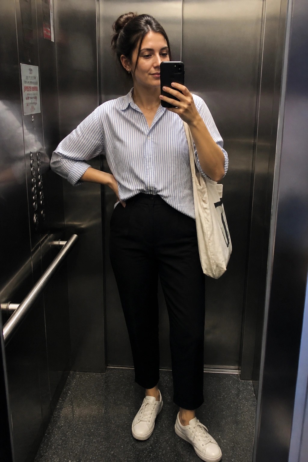 Woman in striped shirt and black pants taking selfie in elevator with tote bag
