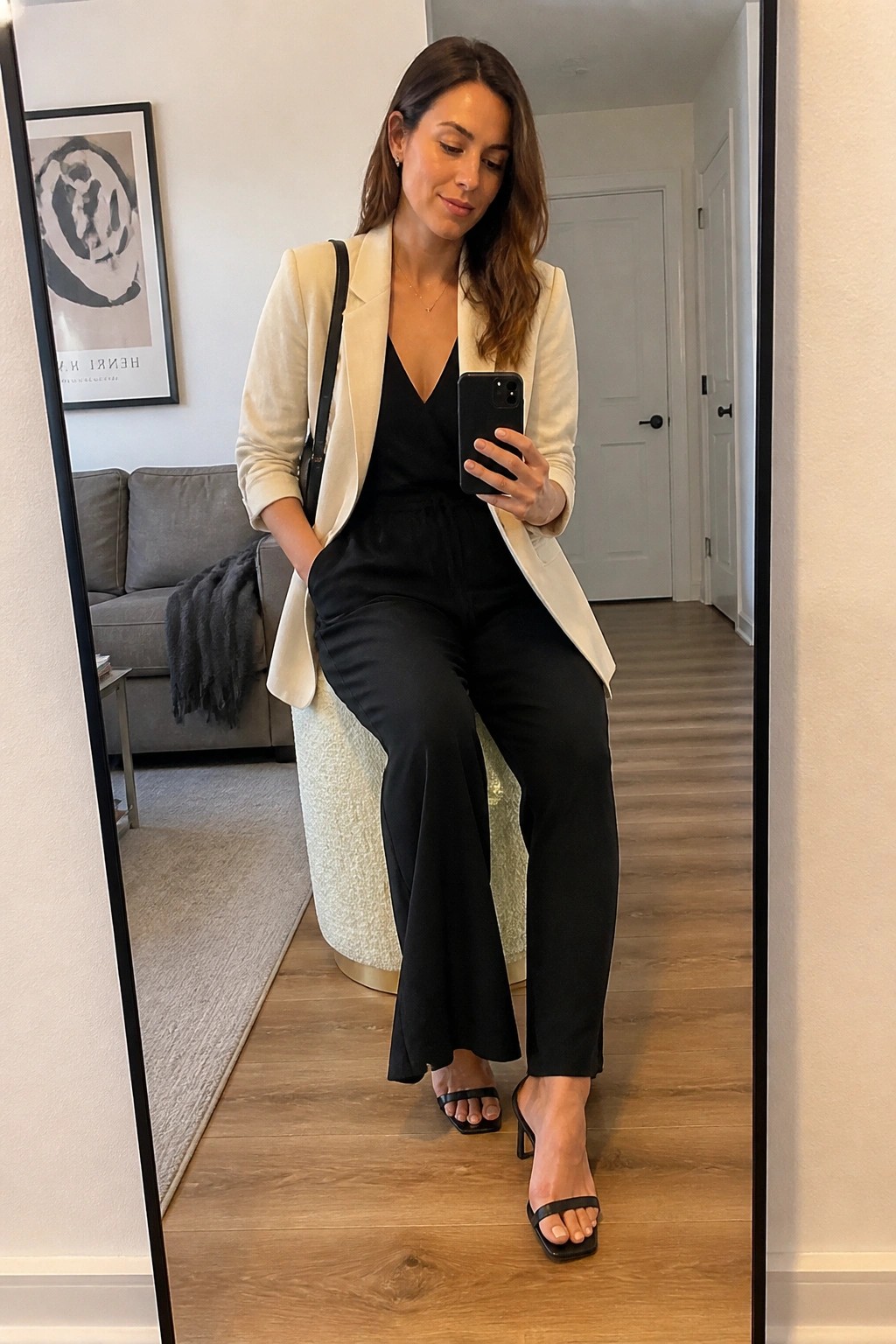 Woman in cream blazer and black jumpsuit taking mirror selfie with phone