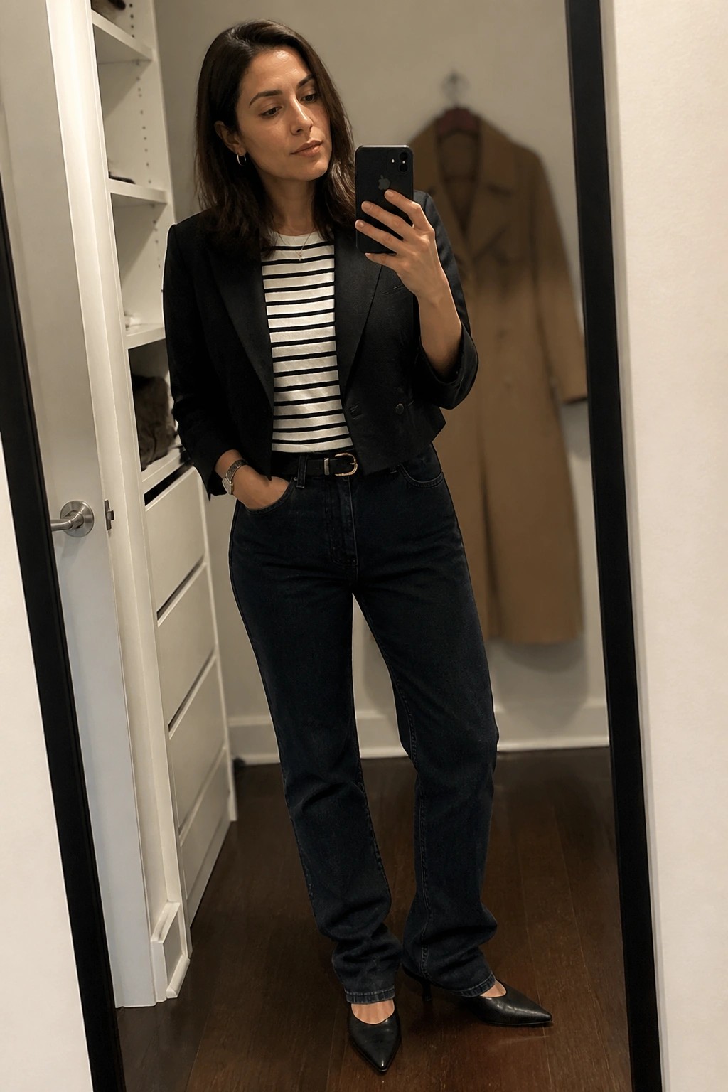 Woman in black blazer and striped top taking mirror selfie in jeans and heels