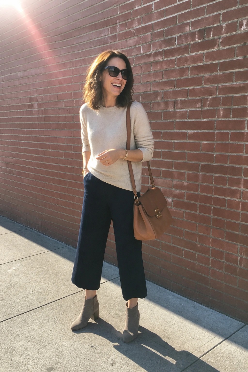 Woman in beige sweater and navy pants smiling by brick wall with brown bag