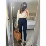 Woman in cream blouse and black pants holding brown bag in bedroom mirror selfie