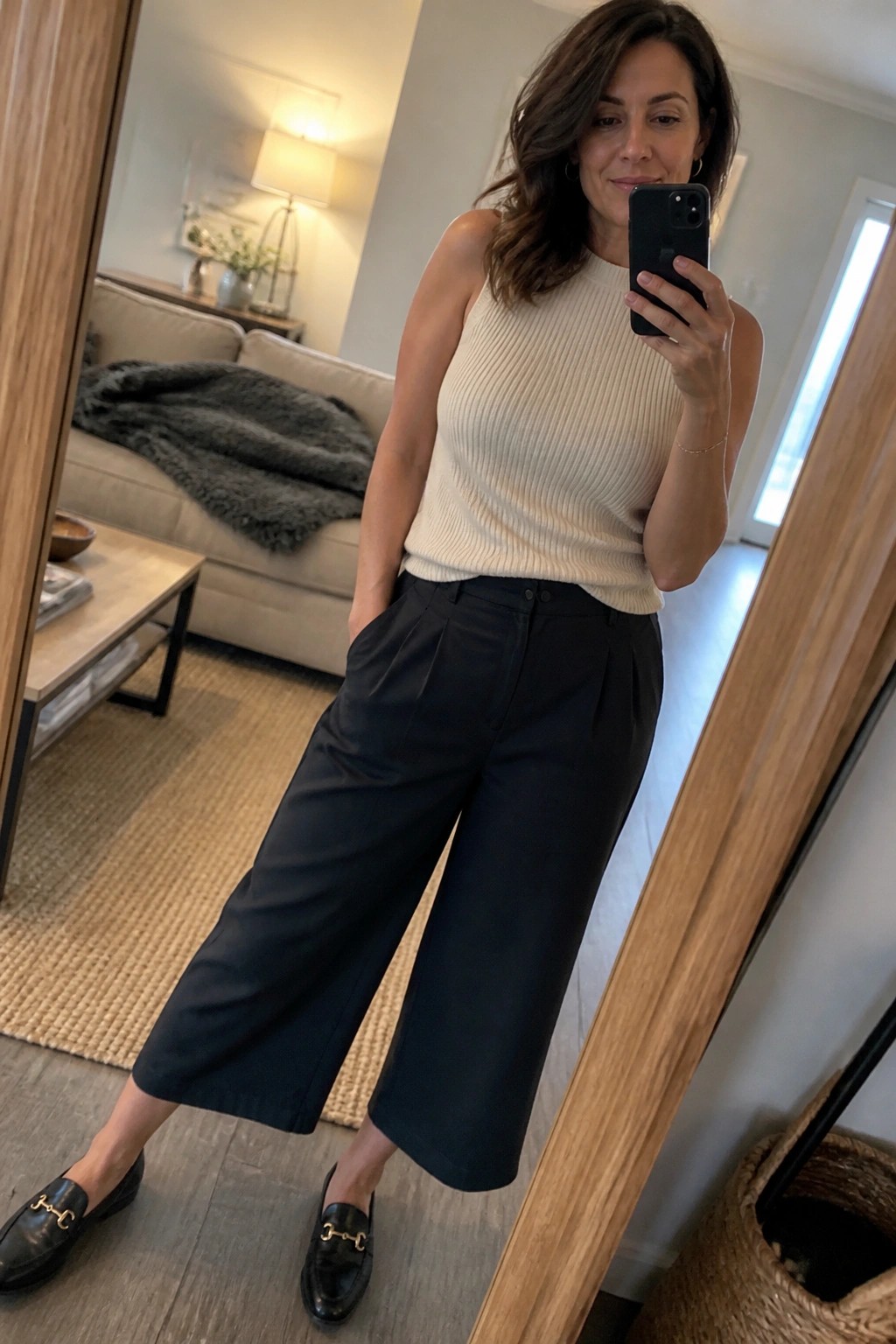 Woman in a beige ribbed sleeveless knit tank tucked into black pleated wide-leg cropped trousers with black loafers featuring gold hardware, taking a mirror selfie in a living room setting.