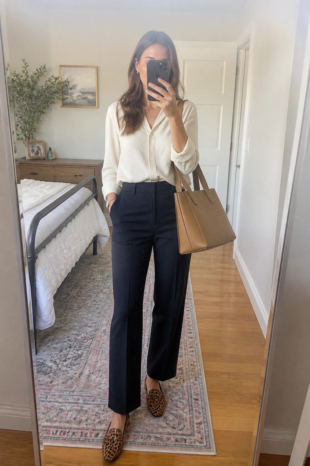 Woman in cream blouse and black pants holding brown bag in bedroom mirror selfie