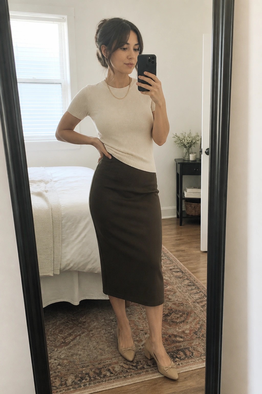 Woman in cream top and brown midi skirt taking bedroom mirror selfie with phone
