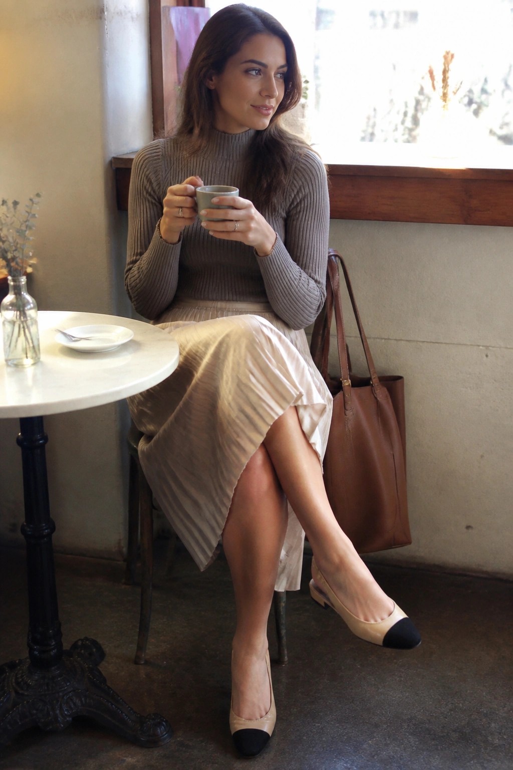 Woman in gray turtleneck and beige skirt holding mug at cafe table