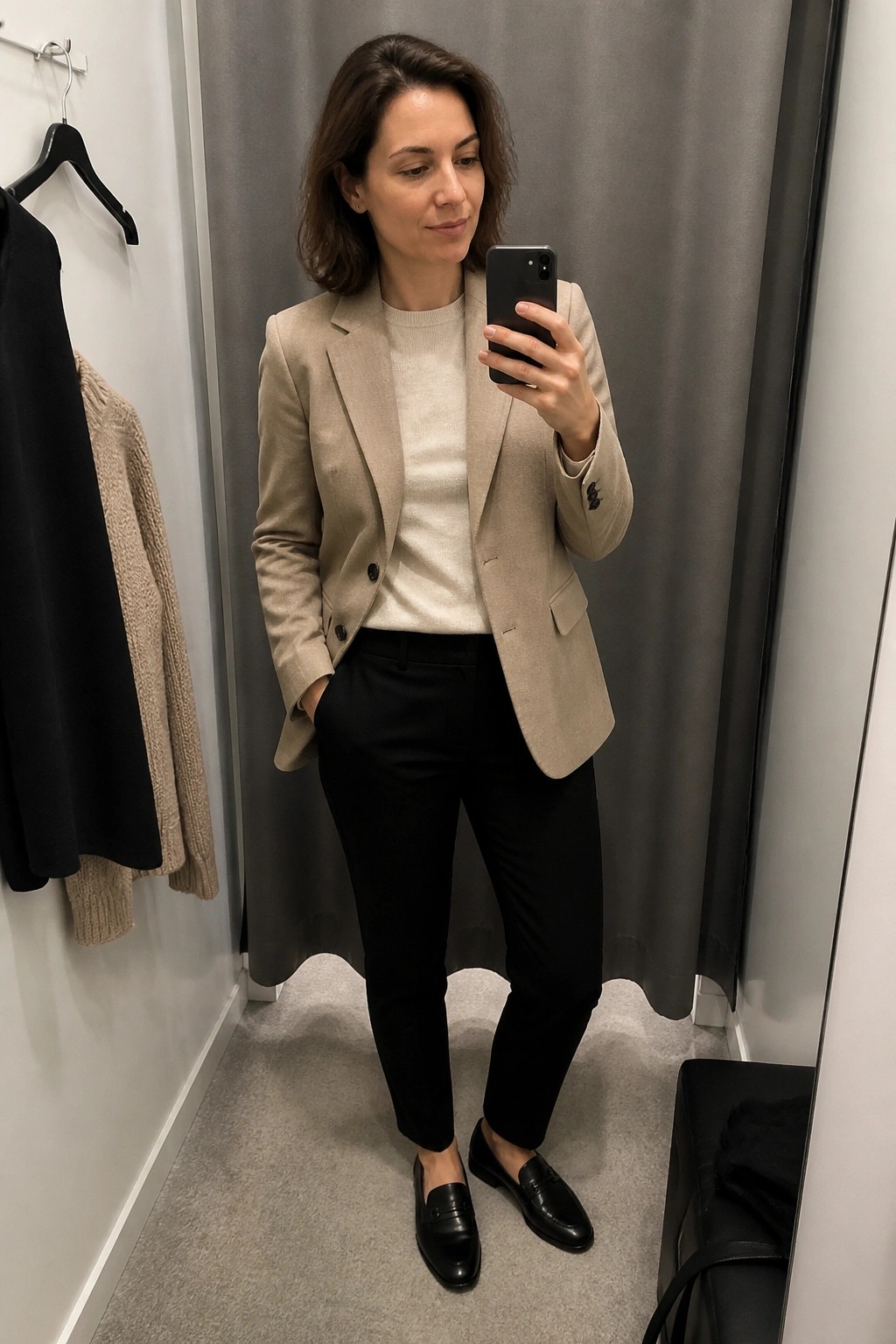 Woman in beige blazer and black pants taking mirror selfie in fitting room