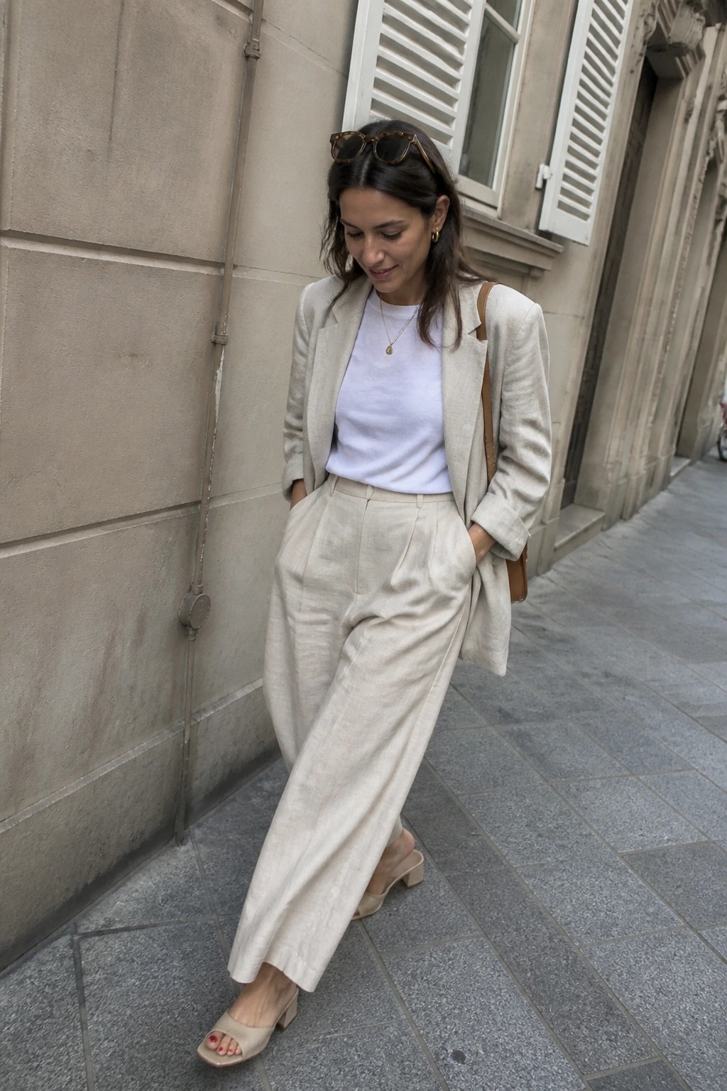 Woman wearing a beige linen blazer over a white t-shirt with matching wide-leg linen trousers, beige heeled mules, and a delicate gold necklace.