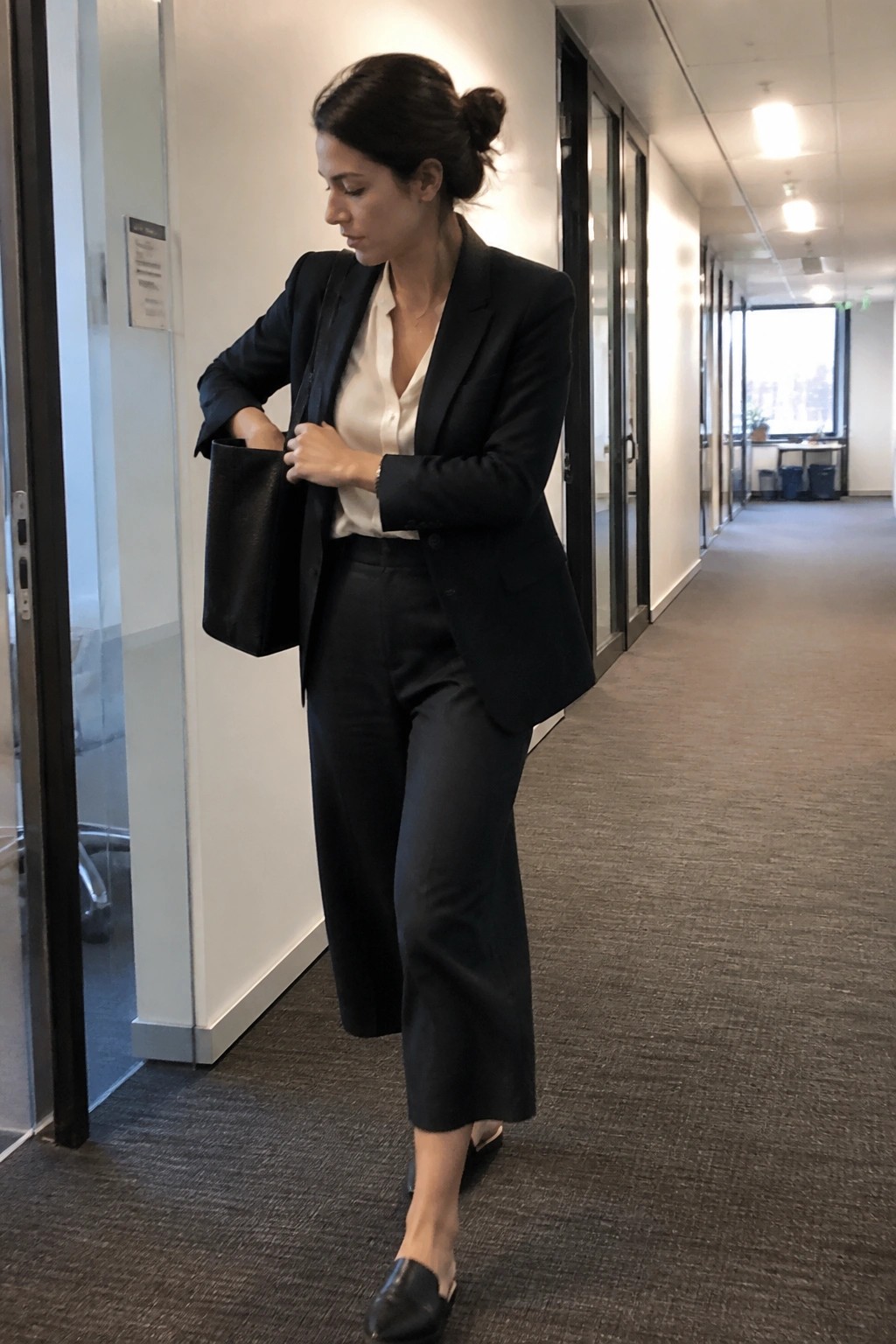 Woman in black blazer adjusting tote bag in modern office hallway