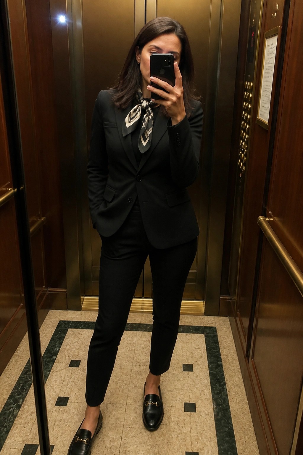 Woman in black pantsuit and scarf taking mirror selfie in elevator