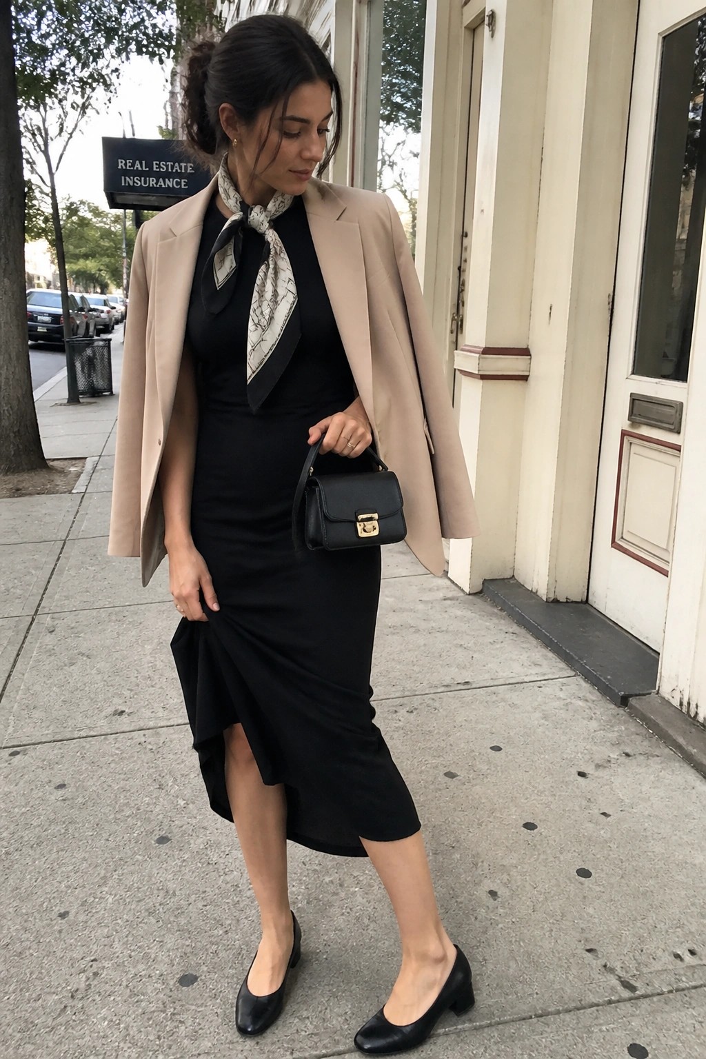 Woman in beige blazer over black midi dress with patterned neck scarf, small black handbag, and black block heel shoes standing on sidewalk.