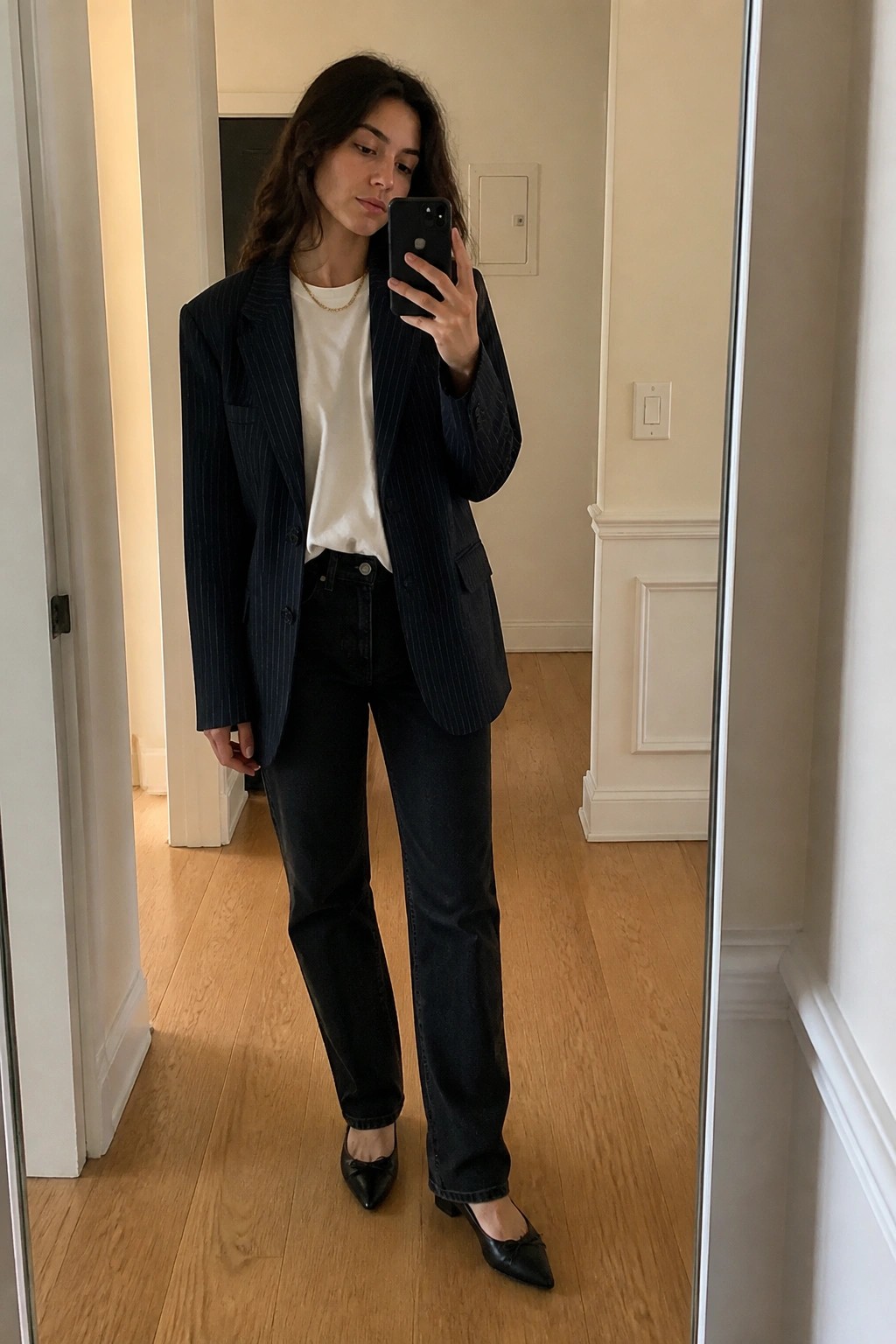 Woman in navy pinstripe blazer over white t-shirt with dark jeans, black pointed flats, and thin gold necklace, taking a mirror selfie.