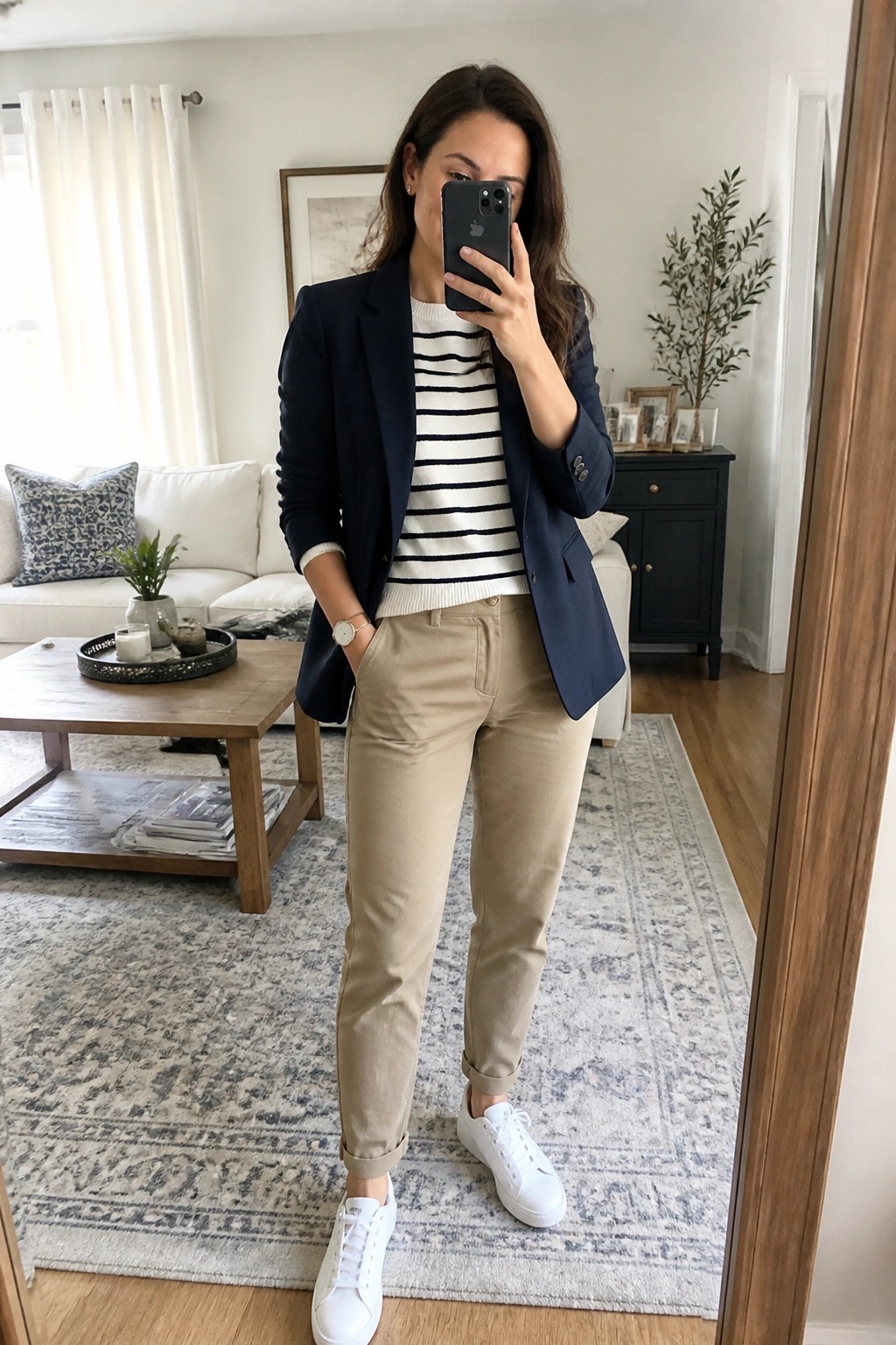 Woman wearing navy blazer over black and white striped sweater with khaki pants and white sneakers posing for mirror selfie