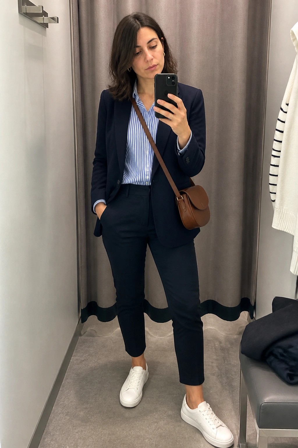 Woman wearing a navy blue pantsuit with a blue and white striped button down shirt, white sneakers, and a brown crossbody bag while taking a mirror selfie in a fitting room.