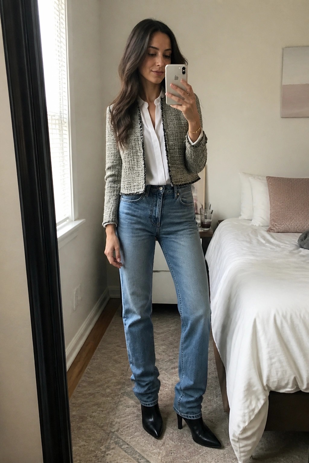 Woman in mirror selfie wearing cropped tweed blazer with black trim over white button down shirt tucked into straight leg blue jeans and black pointed toe heeled boots.