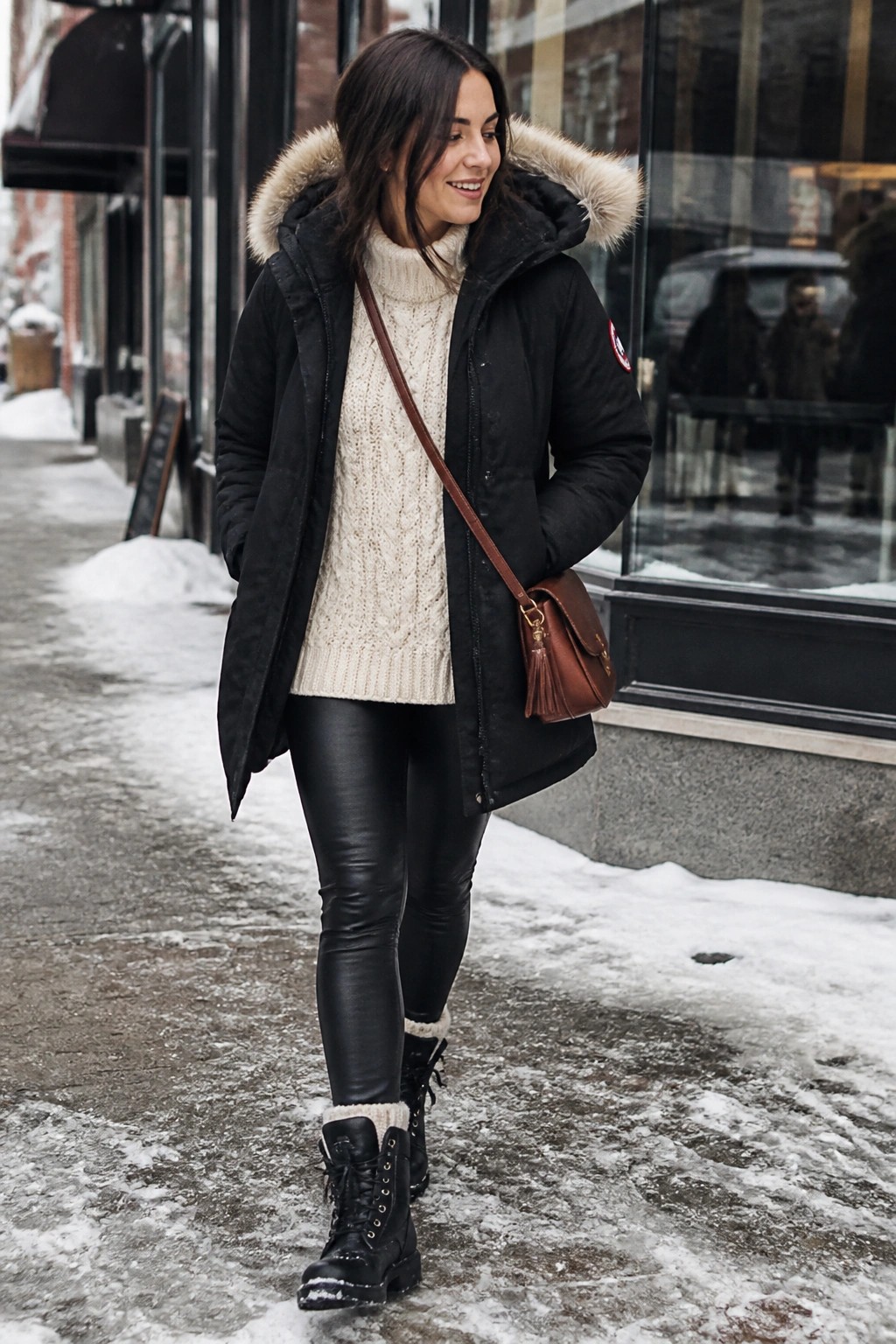 Woman in black fur-hooded parka, cream sweater, and leather pants on snowy street