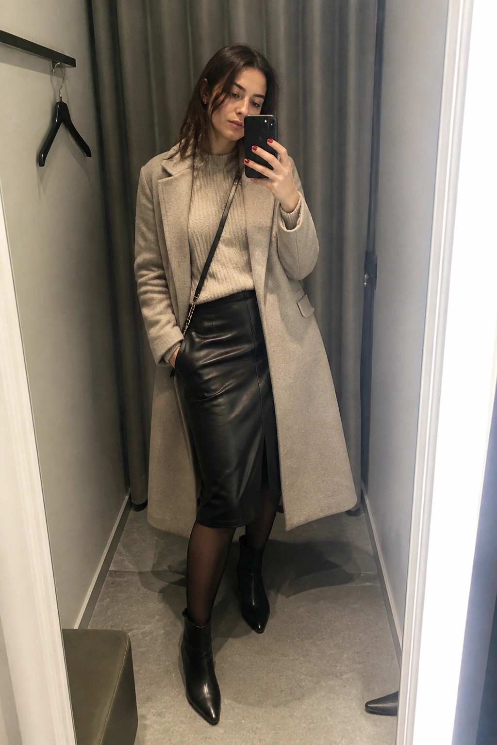 Woman in beige coat and black leather skirt taking mirror selfie in fitting room.