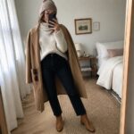 Woman in cream turtleneck, camel coat, jeans, and beanie taking mirror selfie in bedroom