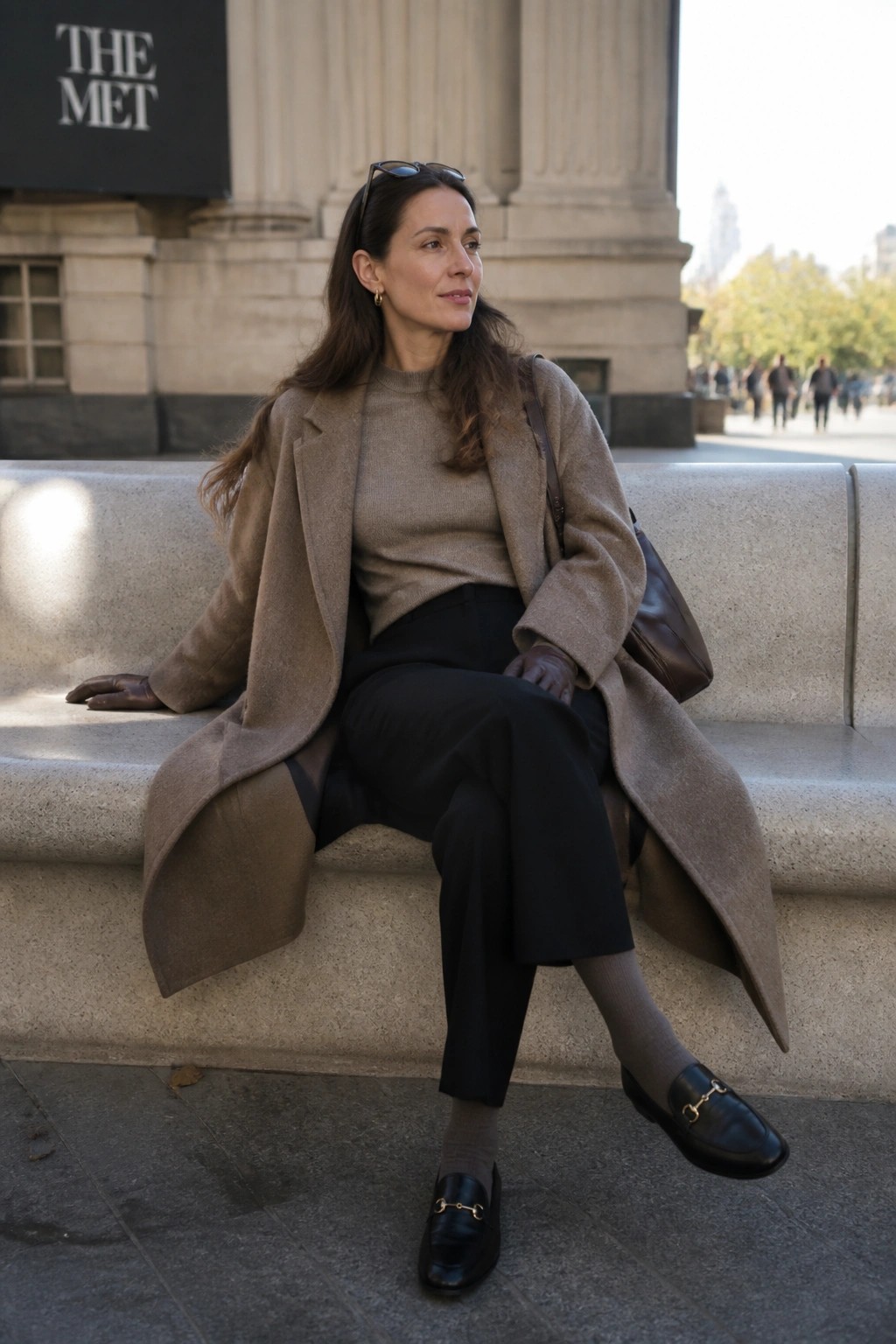 Woman in beige coat and black pants seated on stone bench at The Met